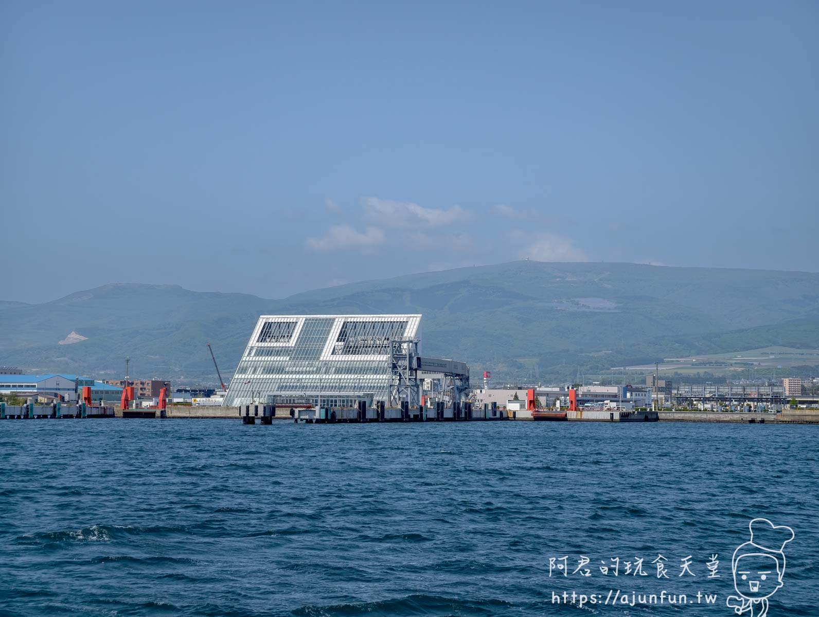 【函館景點】 Bluemoon 港內遊覽船攻略 2026|怎麼搭、票價多少、坐哪個位子才划算 - 第23張圖 函館港外玻璃帷幕地標建築 斜切方形格框結構連接碼頭 函館山脈輪廓 湛藍海水 - 阿君的玩食天堂