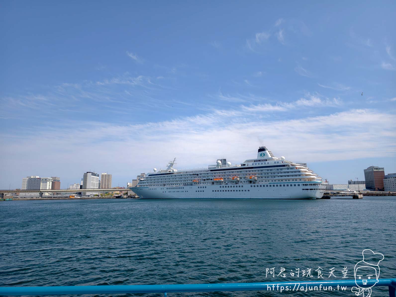 【函館景點】 Bluemoon 港內遊覽船攻略 2026|怎麼搭、票價多少、坐哪個位子才划算 - 第26張圖 函館港大型郵輪停靠 白色多層甲板遠洋郵輪 金色標誌徽章 藍天白雲 Bluemoon視角 - 阿君的玩食天堂