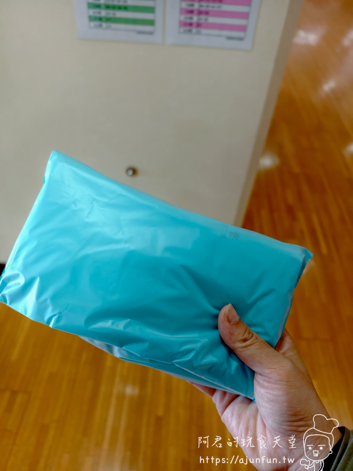 谷地頭溫泉 手持淺藍色塑膠包裝（背景木地板與牆上彩色表格告示）- 阿君的玩食天堂